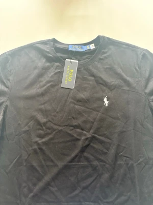 Svart t-shirt från Polo Ralph Lauren - Klassisk svart t-shirt från Polo Ralph Lauren med rund hals och den ikoniska vita logobroderingen på bröstet. Tillverkad i mjuk bomull för en skön känsla och perfekt passform. En stilren basic som funkar till allt.