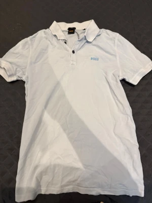 Vit kortärmad polotopp från BOSS - Säljer en vit polotopp från BOSS med klassisk krage och två knappar framtill. T-shirten har korta ärmar och diskret BOSS-logga på bröstet. Perfekt för dig som gillar stilrena och enkla plagg.
