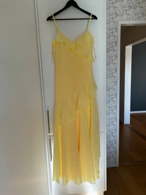 Lång gul balklänning med volanger - Säljer en lång gul klänning med tunna axelband och v-ringad hals. Klänningen har volangdetaljer både upptill och längs kjolen.
