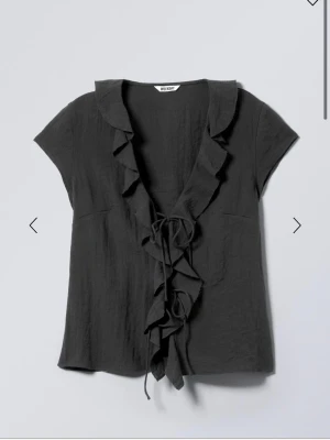 Svart blus med volang från Weekday - Säljer en svart blus från Weekday med volangkant längs v-ringningen och knytband framtill. Blusen har korta ärmar och är tillverkad i ett lätt, skrynkligt material som ger en avslappnad look. Perfekt för att skapa en trendig och cool stil.