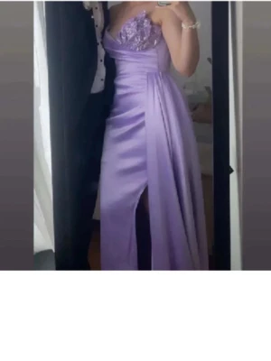 Lila långklänning med spets och slits - Elegant lila långklänning med spetsdetaljer på bröstet och en djup slits framtill. Klänningen har omlott-effekt och är axelbandslös, perfekt för bal eller fest. Materialet har en satinliknande finish som ger en lyxig känsla.