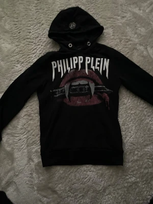 Svart hoodie från Philipp Plein - Cool svart hoodie från Philipp Plein med vampyrtema. Stora vita logotypen och grafiskt tryck med vampyrtänder och blod på både fram- och baksidan. Huva med metallmärke framtill. Perfekt för dig som gillar edgy streetstyle.
