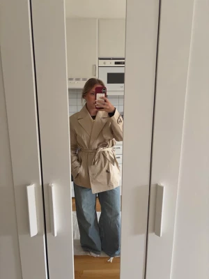 Beige trenchcoat med knytskärp - Stilren beige trenchcoat med breda slag och knytskärp i midjan. Jackan har klassiska detaljer som dubbelknäppning och sidofickor. Från H&M och i storlek S.