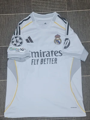 Real Madrid matchtröja Adidas vit - Snygg vit Real Madrid matchtröja från Adidas med klubbmärke på bröstet, gråa och gula detaljer samt svarta ränder på axlarna. Tryck med 'Emirates Fly Better' på framsidan och UEFA Foundation-märke på ärmen. Tillverkad i lätt och ventilerande material.