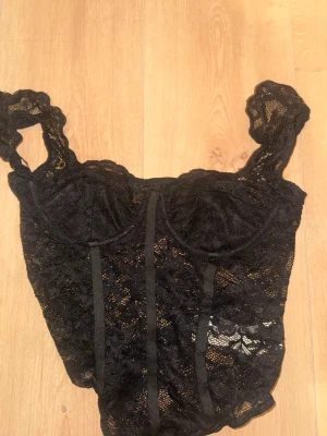 Svart spetsbody med breda axelband - Snygg svart body i genombruten spets med breda axelband och markerade kupor. Bodyn har dekorativa sömmar framtill och är tillverkad i ett stretchigt, transparent material som ger en sexig look. Perfekt att styla under en kavaj eller till jeans.