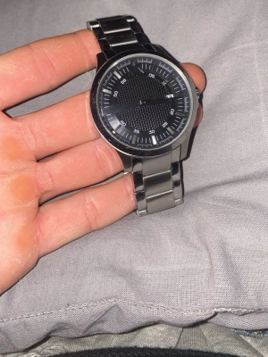 Svart herrklocka från Armani Exchange - Stilren herrklocka från Armani Exchange med svart urtavla och datumvisning. Klockan har ett klassiskt armband i silverfärgad metall och tydliga vita markeringar på urtavlan. Perfekt accessoar för dig som gillar en modern och tidlös look.