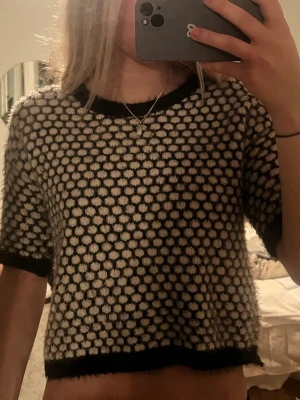Svartvit stickad topp med prickmönster - Säljer en kortärmad stickad topp i svart och vitt med ett coolt prickmönster. Tröjan har svarta ribbade kanter vid ärmslut, halsringning och nederkant. Materialet ser fluffigt och mjukt ut, perfekt för en trendig look. 