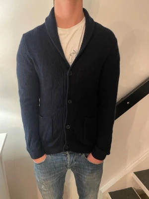 Stickad kofta från Dressmann - Säljer en mörkblå stickad kofta med sjalkrage och knäppning framtill. Koftan har två framfickor och ribbade muddar vid ärmslut och nederkant. Perfekt för lager på lager och enkel att matcha med jeans eller t-shirt. Nypris 600 kr