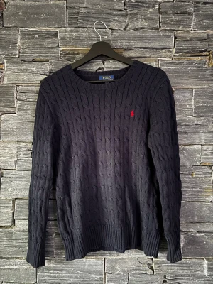 Mörblå kabelstickad tröja från Polo Ralph Lauren - Kabelstickad mörkblå tröja från Polo Ralph Lauren med rund hals och lång ärm. Tröjan har ett diskret rött broderat logotyp på bröstet och ribbade muddar vid ärmslut och nederkant. Perfekt för dig som gillar klassisk och stilren design.
