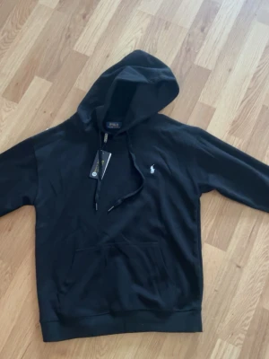 Svart hoodie från Polo Ralphh Lauren - Snygg svart hoodie från Polo Ralph Lauren med klassisk huva och dragsnören. Har en vit broderad logga på bröstet och en stor magficka framtill. Tillverkad i mjukt bomullsmaterial som är skönt att bära. Perfekt för en avslappnad och stilren look.