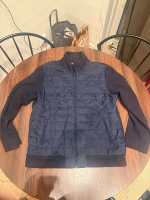 Mörkblå quiltad cardiganjacka - Snygg mörkblå cardiganjacka med quiltad front och mjuka ärmar. Jackan har dragkedja framtill, två fickor med dragkedja och ribbade muddar. Perfekt för lager på lager och en clean look.