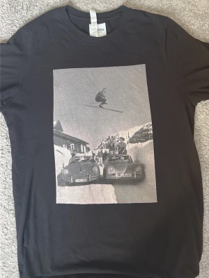 Svart t-shirt med retro print Swedish - Svart t-shirt från Swedish med ett stort retroinspirerat print framtill som visar en skidåkare som hoppar över två klassiska bilar i ett snöigt landskap. T-shirten har rund hals och korta ärmar. Perfekt för dig som gillar unik streetwear. Storlek M passar till storlek S också