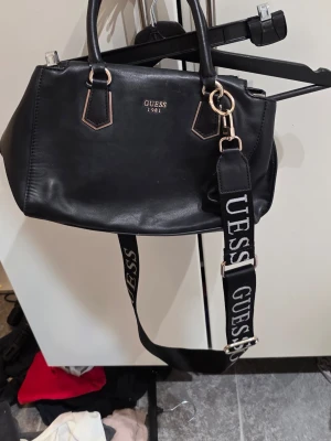 Svart handväska från Guess - Snygg svart handväska från Guess med guldfärgade detaljer och logotypen 'Guess 1981' framtill. Väskan har både handtag och en bred axelrem med Guess-tryck i vitt. Tillverkad i konstläder och har en stilren, boxig form.
