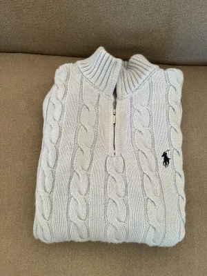 Ljusblå kabelstickad tröja Polo Ralph Lauren - Snygg ljusblå kabelstickad tröja från Polo Ralph Lauren med halv dragkedja och ribbad krage. Tröjan har det klassiska broderade logotypen på bröstet och är gjord i mjukt stickat material. Perfekt för dig som gillar stilrena och tidlösa plagg.