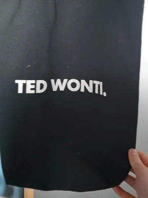Svart tygkasse från Ted Wonti - Snygg svart shoppingväska i tyg från Ted Wonti med vit text på framsidan. Perfekt för att bära böcker, laptop eller annat du behöver under dagen. Enkel och stilren design med rejäla handtag.