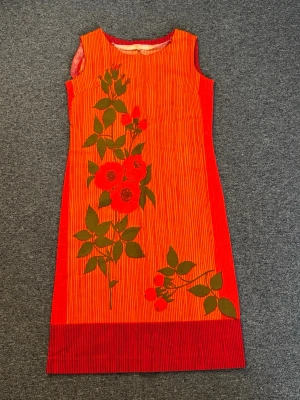 VINTAGE - 60 talet. Orange-randig blommig ärmlös klänning - Vintage orange klänning från 60 talet med tunna vertikala ränder och stora blommiga tryck i grönt och rött framtill. Klänningen är ärmlös och har en rak passform med bred röd kant längst ner. Perfekt för dig som vill sticka ut med färg och mönster. Eller som älskar äkta vintage stil:) oanvänd och utan defekter, fläckarna är bara damm då den inte kommit till användning 