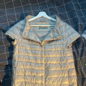 Beige glansig dunväst från Herno - Snygg beige glansig dunväst från Herno med korta ärmar och hög krage. Västen har quiltad design och tryckknappar vid halsen. Perfekt för lager på lager och ger en trendig look till din outfit.