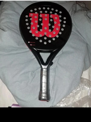 Wilson padelracket svart/röd - Säljer ett padelracket från Wilson i svart med stor röd logga på träffytan. Racketen har rund form, många hål och grepp i svart material. Perfekt för dig som vill ta ditt padelspel till nästa nivå.