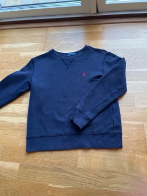 Mörkblå sweatshirt från Polo Ralph Lauren - Klassisk mörkblå sweatshirt från Polo Ralph Lauren med rund halsringning och röd broderad logga på bröstet. Tröjan har långa ärmar och ribbade muddar vid ärmslut och nederkant. Perfekt för en avslappnad och stilren look. Storlek M för barn ca 10-12 år