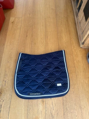 Marinblått schabrak Copperfield STL: FULL DRESSYR - Snyggt marinblått schabrak från Copperfield med quiltat mönster och ljusblå kantband. Tillverkat i ett glansigt material som ger en exklusiv känsla. Perfekt till hästutrustningen för dig som vill ha stilrena detaljer.
