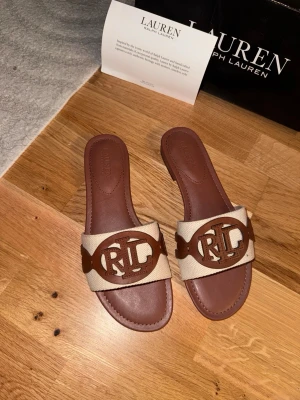Bruna sandaler från Lauren Ralph Lauren - Säljer ett par stilrena sandaler från Lauren Ralph Lauren i brunt och beige. Sandalerna har en bred rem i textil med ett stort RL-logotyp i brunt läder framtill. Platt sula och öppen tå ger en avslappnad look. Perfekta för sommaren och lätta att matcha.