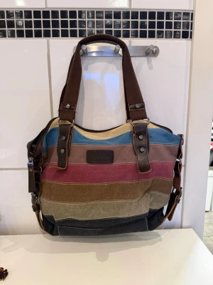 handväska med bruna detaljer - Säljer en färgglad handväska med breda ränder i blått, rosa, brunt, beige och svart. Väskan har bruna handtag och detaljer i konstläder samt en praktisk dragkedja och ytterfack. Perfekt för dig som gillar en avslappnad och unik stil. Perfekt för dig som gillar en Vinted look.