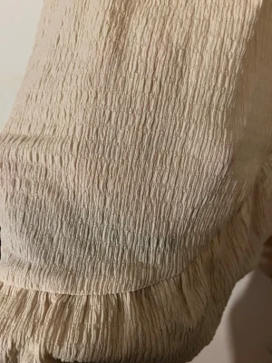 Beige omlottklänning med volang - Säljer en beige omlottklänning med v-ringning och knyt i midjan. Klänningen har korta puffärmar och volangdetaljer längs nederkanten och omlotten. Materialet är strukturerat och känns lätt och luftigt, perfekt för varmare dagar. 