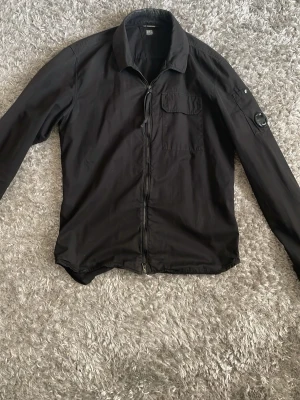Svart overshirt från C.P. Company - Stilren svart overshirt från C.P. Company med dragkedja framtill, bröstficka och ikonisk linsdetalj på ärmen. Tillverkad i ett lätt och bekvämt material, perfekt för lager-på-lager. Klassisk krage och långa ärmar med knapp vid ärmslut. XS-S. Kan gå ner i pris vid snabb och smidig affär.