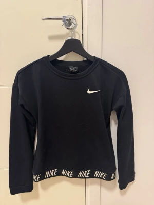 Svart Nike Dri-Fit tröja - Svart långärmad tröja från Nike med Dri-Fit material. Tröjan har rund halsringning, vit Nike-logga på bröstet och elastisk nederkant med upprepat Nike-tryck. Perfekt för träning eller chill. Passar dig som är 12-13 år eller 146-156 cm.