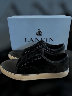 Svarta lanvins - Snygga svarta sneakers från Lanvin med ovandel i mocka och detaljer i blankt läder. Skorna har vita gummisulor och klassisk låg profil med svarta skosnören och metallöljetter. Perfekta för en clean och stilren look.