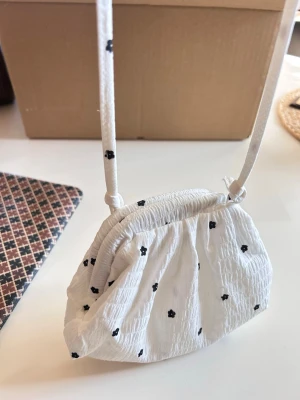 Vit handväska med små svarta blommor - Söt vit handväska i tyg med små svarta blommor och rynkad design. Väskan har en tunn axelrem och stängs med dragsko. Perfekt för dig som gillar en gullig och enkel stil. Materialet känns lätt och väskan har en mjuk, puffig form.