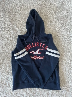 Mörkblå Hollister hoodie med tryck - Snygg mörkblå hoodie från Hollister med stor logga och texten 'California' i rött och vitt på bröstet. Hoodien har huva med dragsko, vita ränder på ärmarna och en klassisk känguruficka framtill. Perfekt för en avslappnad och sportig stil.