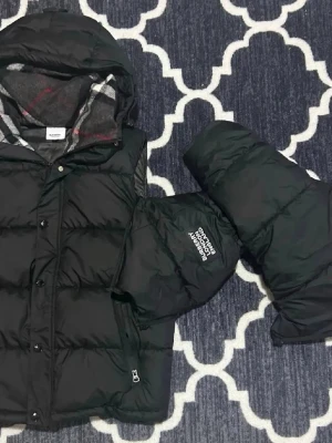 Burberry jacka, priset diskutera  - Priset går att diskutera . Stilren svart dunväst från Burberry med huva och klassiskt rutigt innerfoder. Västen har tryckknappar, dragkedja och Burberry-logga på axeln. Perfekt för lager-på-lager och håller dig varm under kyliga dagar.