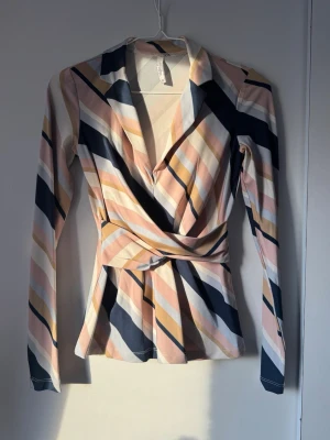 Randig omlottblus i pastellfärger - Säljer en snygg omlottblus med långa ärmar och krage. Blusen har diagonala ränder i rosa, beige, marinblått och vitt. Materialet känns mjukt och stretchigt, perfekt för en trendig look. Sitter figurnära med markerad midja och omlottdetalj framtill.