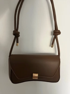 Brun axelväska med gulddetalj - Snygg brun axelväska med axelband och knutdetaljer. Väskan har ett guldfärgat spänne framtill. Perfekt storlek för det viktigaste och stilren design som passar till många outfits. Aldrig använd, så mycket bra skick