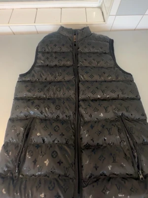 Svart dunväst från Louis Vuitton - Snygg svart dunväst från Louis Vuitton med ikoniskt monogrammönster över hela västen. Västen har hög krage, dragkedja framtill och två praktiska fickor med dragkedja. Materialet är glansigt och ger en exklusiv känsla.