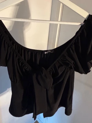 Svart offshoulder topp från Gina Tricot - Trendig svart offshoulder topp från Gina Tricot med korta ärmar och en snygg knytning framtill. Toppen har en croppad passform och är perfekt för dig som vill ha en stilren men ändå cool look. Tillverkad i mjukt material som känns skönt mot huden.