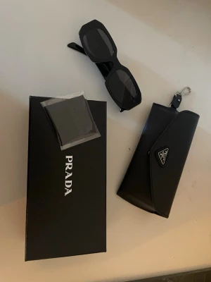 Svarta solglasögon från Prada - Snygga svarta solglasögon från Prada med rektangulära bågar och klassisk logga på skalmen. Kommer med originalfodral, putsduk och box. Perfekta för dig som vill ha en stilren och exklusiv accessoar till sommaren.