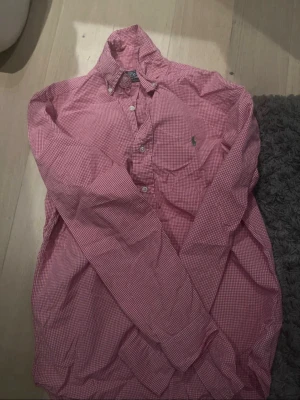Rutig rosa skjorta från Ralph Lauren - Klassisk långärmad skjorta från Ralph Lauren i rosa och vitt rutigt mönster. Skjortan har knappar framtill och den ikoniska gröna loggan broderad på bröstet. Tillverkad i bomull och har en tidlös, normal passform.