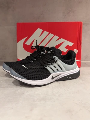 Nike Air Presto svart/vit sneakers - Nike Air Presto sneakers i svart mesh med vita och grå detaljer. Skorna har en låg silhuett, vit sula och svart swoosh-logga på sidan och tån. Snygg design med plastdetaljer vid snörningen och bekväm passform för en sportig look.
