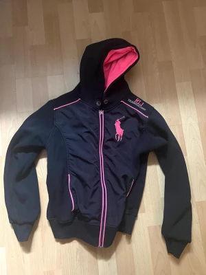 Svart hoodie RL1 Team Racing - Snygg svart hoodie / kofta med dragkedja från Ralph lauren Team Racing. Hoodien har rosa detaljer, inklusive dragkedja, sömmar och stor logga på bröstet. Insidan av huvan är rosa vilket ger en cool kontrast. Perfekt för dig som gillar sportig stil och vill sticka ut. Har en fläck i luvan som jag ej testat att få bort. Går säkert bort med fläckrengöring 