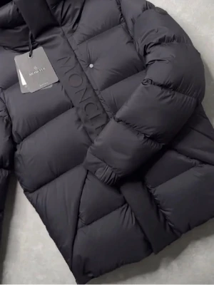 Svart dunjacka från Moncler - Snygg svart dunjacka från Moncler med quiltad design och stora, fluffiga partier. Jackan har hög krage, tryckknappar och Moncler-logga längs dragkedjan. Perfekt för kalla vinterdagar och riktigt trendig stil.