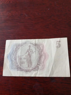 Svensk femkronorssedel 1960 - Femkronorssedel från 1960 utgiven av Sveriges Riksbank. Sedeln har ett klassiskt motiv med kungaporträtt på framsidan och en kvinnofigur med sköld på baksidan. Färgerna är främst bruna och blåaktiga nyanser. Perfekt för samlare av gamla svenska sedlar.