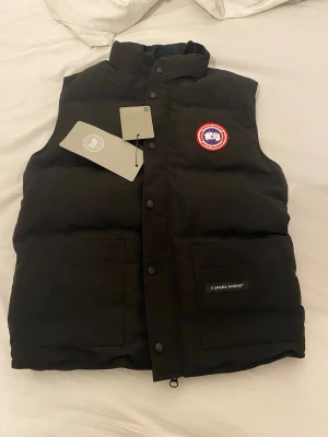Svart dunväst från Canada Goose - Snygg svart dunväst från Canada Goose med hög krage, två stora framfickor och klassisk logotyp på bröstet. Västen har tryckknappar framtill och är fylld med dun för extra värme. Perfekt för lager-på-lager och streetstyle.