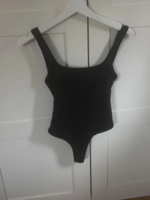 Svart basic body med breda axelband - En enkel och stilren svart body med breda axelband och rundad urringning. Bodyn har en klassisk passform och är tillverkad i ett stretchigt material som sitter bekvämt mot kroppen. Perfekt att matcha med jeans eller kjol för en clean look. Inga defekter förutom att jag klippt av banden som är till för att hänga upp bodyn pga att dom stack ut!