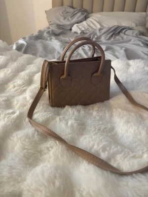 Beige quiltad minihandväska - Säljer en beige minihandväska med quiltat mönster och både handtag och axelrem. Väskan har en stilren, rektangulär form och dragkedja upptill. Perfekt för dig som gillar trendiga accessoarer och vill ha en liten väska till mobilen och det viktigaste.