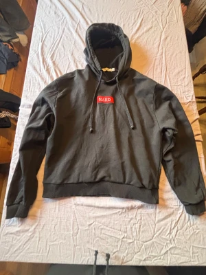 Svart hoodie från NA-KD med logga - Svart hoodie från NA-KD med röd logga på bröstet. Klassisk modell med huva och snörning samt ribbade muddar vid ärmslut och nederkant. Perfekt för en chill och avslappnad stil.