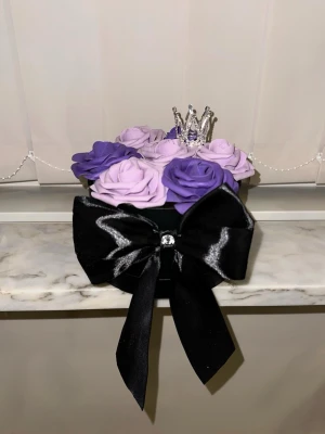 Dekorativ blombox med rosetter och krona - Säljer en rund svart blombox dekorerad med lila och ljuslila konstgjorda rosor, en glittrig silverfärgad krona på toppen och en stor svart sidenrosett framtill. Perfekt som present eller inredningsdetalj.