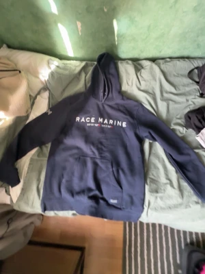 Mörkblå hoodie från Race Marine - Mörkblå hoodie från Race Marine med vit text på bröstet och klassisk känguruficka framtill. Tröjan har huva och ribbade muddar vid ärmslut och nederkant. Perfekt för en avslappnad och sportig stil.