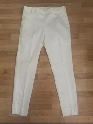 Vita kostymbyxor med pressveck - Snygga vita kostymbyxor med raka ben och klassiskt pressveck. Byxorna har hällor för skärp och sidofickor. Perfekta för en clean och stilren look. Materialet ser ut att vara bomull eller en bomullsblandning. Använd en gång.